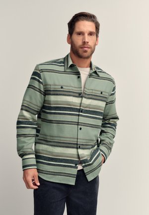 Mand iført en grøn, grå og sort stribet skjorte med knapper over en hvid T-shirt med hænderne i lommerne på mørke bukser, neutral baggrund.