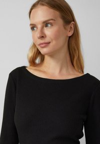Kobieta z długimi blond włosami, ubrana w czarny sweter w prążki, patrzy nieco w bok na tle jasnego, jednolitego tła.
