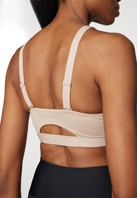 Beige idretts-BH med teksturert overflate, justerbare stropper og utskjæring på baksiden. Materialet ser ut til å være elastisk og støttende.