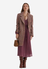 Kvinde iført en lang lilla kjole, ternet frakke, brune støvler, guldkæde og holder en brun pyntet håndtaske mod en hvid baggrund.