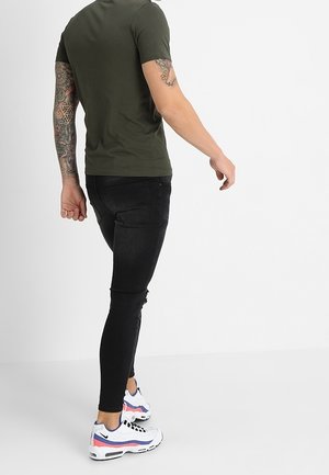 Olivengrøn tætsiddende t-shirt, sorte slim jeans med slidte detaljer og mønstrede hvide atletik sko med pink og blå accenter.