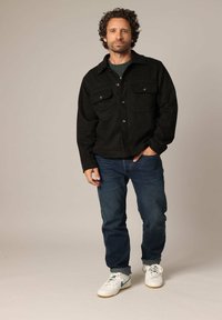 Veste en daim noire avec deux poches poitrine, portée sur une chemise verte, accompagnée de jeans bleus et de baskets blanches avec des accents bleus.