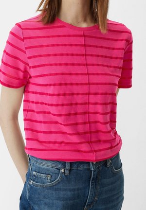 Kobieta ubrana w różowy t-shirt z krótkim rękawem z ciemniejszymi różowymi poziomymi paskami i niebieskie dżinsy na jednolitym tle.