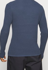 Homme portant un pull ajusté à manches longues en maille gaufrée bleu marine et un pantalon noir, vu de dos sur un fond uni.