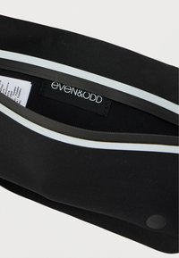 Pochette noire au toucher lisse, ornée d'une bande blanche et d'une quincaillerie ronde noire. L'intérieur présente une étiquette de marque et une étiquette d'entretien.