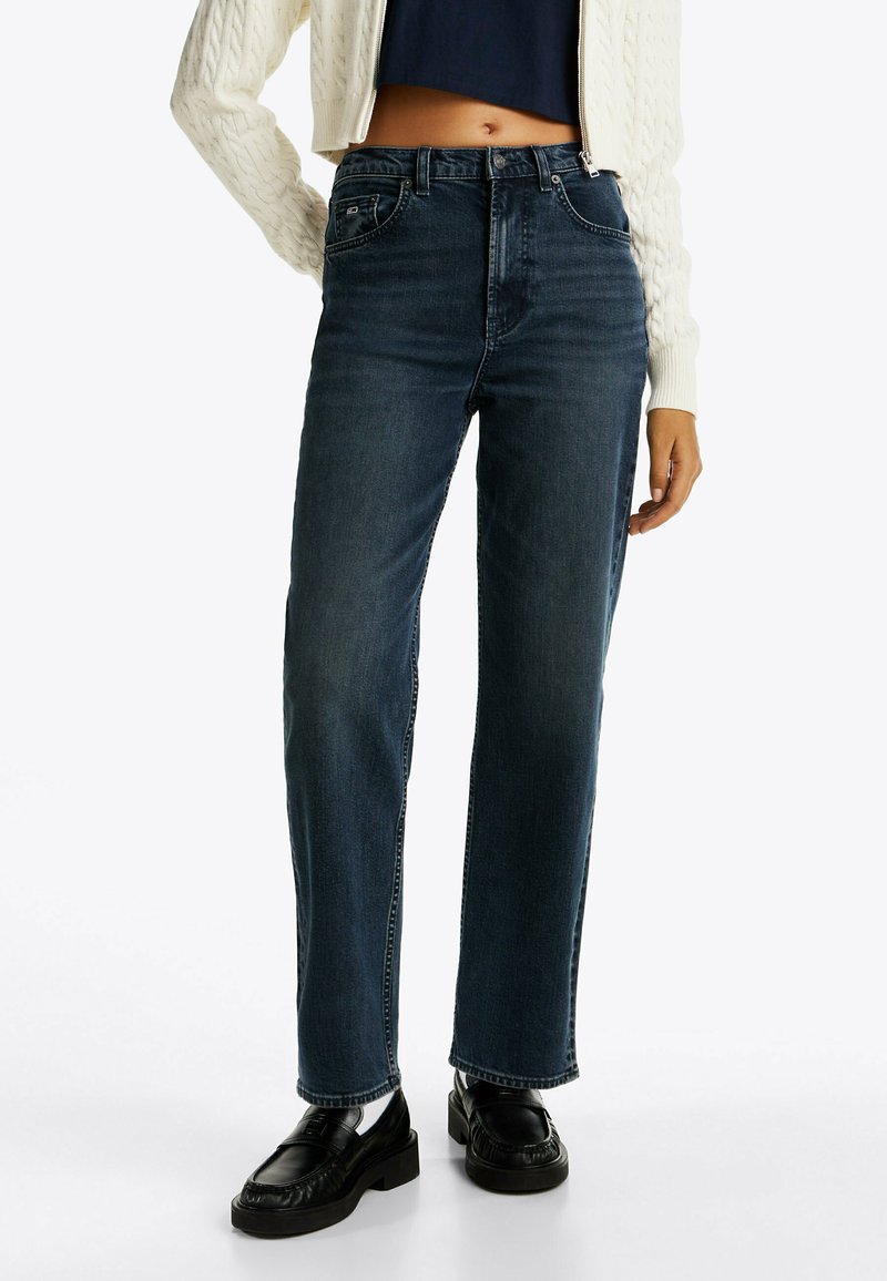 Jeans de denim de lavado oscuro, de pierna recta, que presentan una cintura alta, bolsillos frontales y costuras en contraste, combinados con zapatos negros.