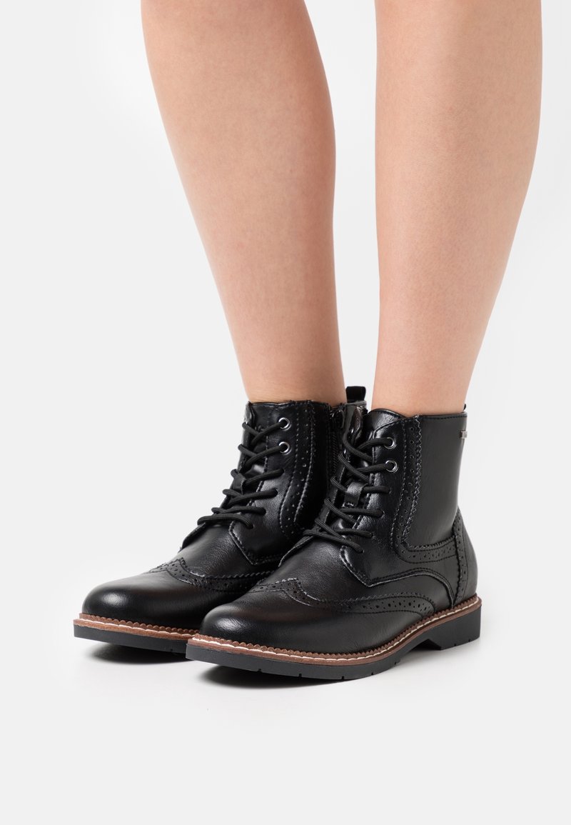 Bottines en cuir noir avec détails brogue, lacets, fermeture éclair latérale et semelle marron cousue, portées sur jambes nues sur fond blanc.