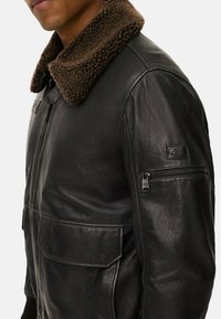Schwarze Lederjacke mit einem braunen Kunstpelzkragen, ausgestattet mit einem Frontreißverschluss, zwei großen geknöpften Taschen und einer Reißverschlusstasche am Ärmel.