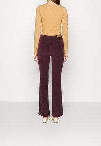 Flared corduroy pants in dark burgundy, featuring back pockets and a waistband label. Paired with a long-sleeve mustard top.
