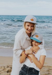 Két ember a tengerparton, azonos szürke sapkával, amelynek türkizszínű karimája van. Az egyik sapkán bőrből készült logós jellemző található. Könnyű szövetű ruházat.