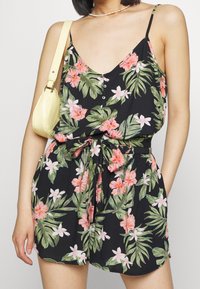 Černý květinový overal s růžovými a zelenými tropickými vzory, s výstřihem do V, zapínáním na knoflíky, nastavovacími ramínky a uvázaným pasem. Akcent v podobě žluté tašky.