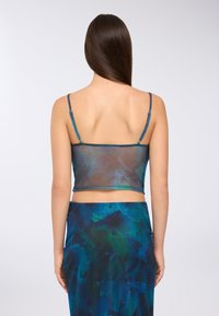 Kobieta z długimi brązowymi włosami, ubrana w przezroczysty niebiesko-zielony top i dopasowaną spódnicę w wzory, odwrócona plecami na jasnym tle.