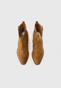 Brūnu zamšādas potītes zābaciņi ar sānu rāvējslēdzējiem un dekoratīvām šuvēm, redzami no augšas uz baltas fona.