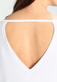 Top bianco con un'apertura a V sul retro. Il tessuto sembra liscio, con una consistenza morbida e spalline sottili sulle spalle.
