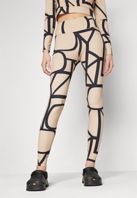 Beige leggings med et svart geometrisk mønster, høy midje og glatt tekstur. Bært med svarte klosser, som viser praktisk fottøy.