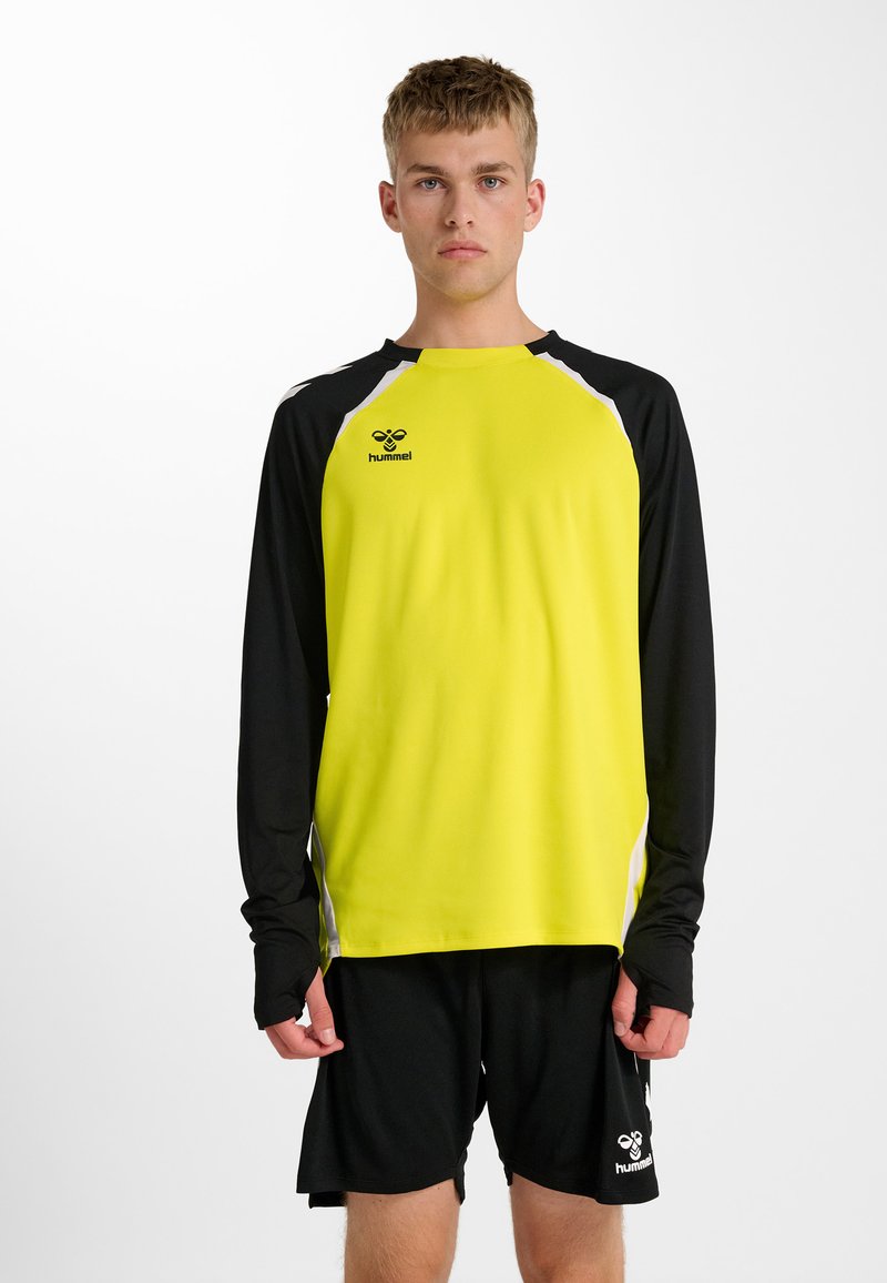 Jonge man die een geel-zwart sportshirt met lange mouwen en een zwarte korte broek met Hummel-logo's draagt, staand tegen een witte achtergrond.