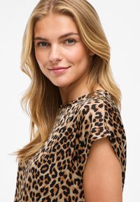 Blouse en imprimé léopard à manches courtes, avec un fond beige et des taches noires/brunes. Tissu doux et léger avec une coupe décontractée.