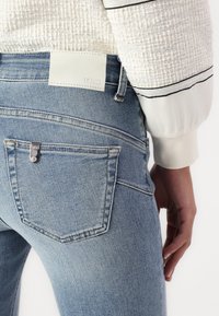 Vaaleansiniset farkut, joissa on hienovarainen tekstuuri, viisi taskua ja valkoinen nahkainen brändilogo vyötäröllä.