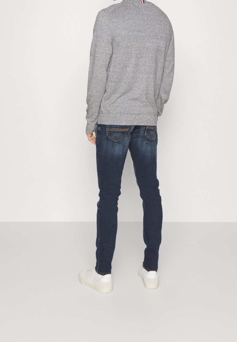 Homme portant un pull gris, un jean bleu foncé et des baskets blanches, debout face à un mur blanc, les mains détendues le long du corps.