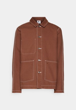 Hnedá button-up bunda Nike s bielym šitím, tromi kovovými gombíkmi, dvoma prednými vreckami a malým vyšívaným logom Nike swoosh na jednom vrecku.