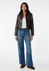 Brun læderjakke med krave, parret med vidde denim jeans i blå. Outfitet har en hvid top og brune sko. Enkel, casual design.