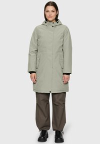 Femme debout portant un parka à capuche gris clair, un pantalon loose marron et des bottes noires devant un fond gris uni.
