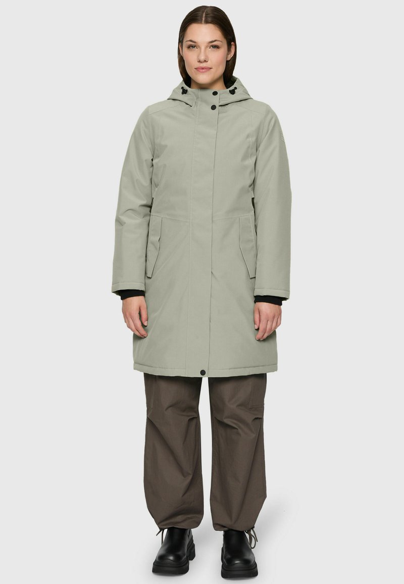 Femme debout portant un parka à capuche gris clair, un pantalon loose marron et des bottes noires devant un fond gris uni.