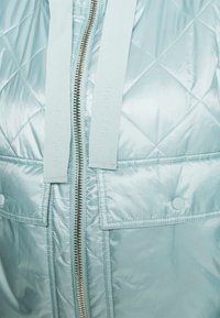 Hellblaues gesteppte Jacke mit Frontreißverschluss, zwei Klappentaschen mit Knöpfen und dicken Zugbändern mit eingeprägtem "Marc O'Polo"-Text.