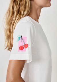 Biała koszulka z grafiką dwóch czerwonych wiśni z zielonymi liśćmi, nadrukowaną na lewym rękawie na różowym tle.