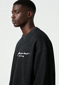 Jonge man in een zwart sweatshirt met de tekst "follow your heart" en een pijl, kijkend naar links tegen een effen lichte achtergrond.