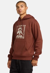 Hoodie marron avec une poche frontale, arborant un design graphique blanc d'une montagne et d'un soleil. Le tissu est doux et légèrement texturé.