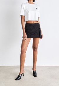 Bílé zkrácené tričko s krátkým rukávem, spárované s černou mini sukni s cargo kapsami a hladkou texturou. Černé podpatkové muly.