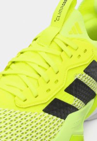 Αθλητικό παπούτσι νέον κίτρινου χρώματος με επάνω μέρος από πλέγμα, υφασμάτινες λεπτομέρειες, μαύρες ρίγες και μαλακή σόλα. Περιλαμβάνει κίτρινη θηλιά στο γλώσσα.