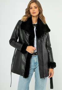 Giacca in pelle nera con un grande colletto e polsini in pelliccia artificiale, dotata di chiusura con zip e vita regolata da una cintura. Texture liscia e vestibilità sartoriale.