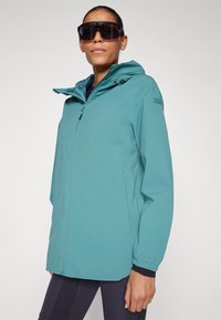 Lettvekts regnjakke i teal med hette, glidelås, elastiske mansjetter og sidelommer. Glatt tekstur med et minimalistisk design.