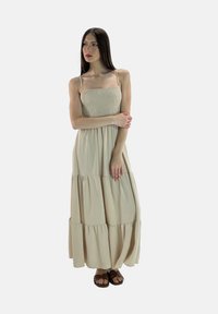 Maxi abito in beige chiaro, caratterizzato da un corpino arricciato, spalline regolabili e gonna a strati. Tessuto morbido con una silhouette fluida.