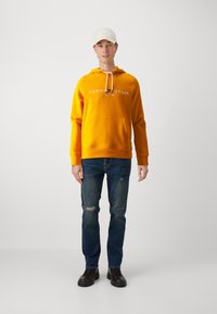 Žltá mikina s kapucňou a bielym logom "Tommy Hilfiger", klokanie vrecko, v kombinácii s tmavomodrými roztrhnutými džínsy a čiernymi šnurovacími topánkami.