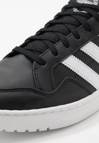 Svart lädersneaker med vit tre-randsdesign, texturerad yta, rund tå och gummisulor. Har svarta snören och sömnadsdetaljer.