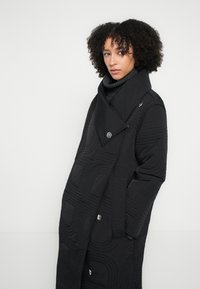 Black quilted coat featuring a high collar, angular lapels, and large silver buttons. The pattern includes raised geometric designs.