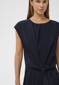 Salopetă bleumarin, cu mânecă scurtă și decolteu rotund, având un front drapat și detalii cu o talie legată. Material neted cu o strălucire subtilă.