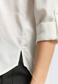 Chemise blanche en tissu tissé avec un motif à petits carreaux subtil, manches retroussables et ourlet arrondi. Associée à un pantalon en tissu noir.