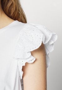 White short-sleeve top with eyelet lace ruffle detailing on shoulder, worn by person with light skin and light brown hair partially visible.