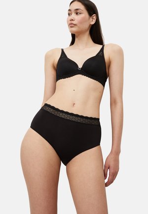 Frau trägt schwarzen BH mit Spitzenbesatz und hoch taillierte Unterwäsche, steht mit seitlich gedrehtem Kopf vor schlichtem Hintergrund.