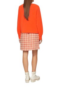 Oranje sweatshirt met lange mouwen, gecombineerd met een gemusterde rok tot de knie in rood en wit hounds-tooth. Witte enkellaarsjes.