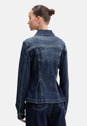 Donna con i capelli raccolti in uno chignon, indossa una giacca di jeans attillata blu scuro e jeans abbinati, vista da dietro contro uno sfondo semplice.
