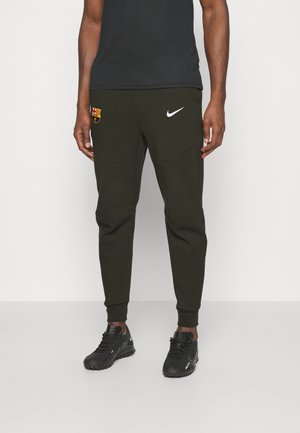 Personne portant un pantalon de sport noir avec les logos Nike et FC Barcelone, des baskets noires et un t-shirt noir, debout sur un fond blanc.