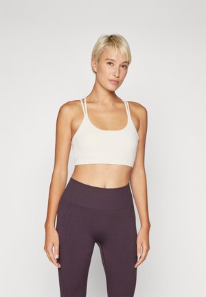 Light beige sports bra with a cropped design, thin straps, smooth texture, paired with high-waisted dark purple leggings featuring seam details.