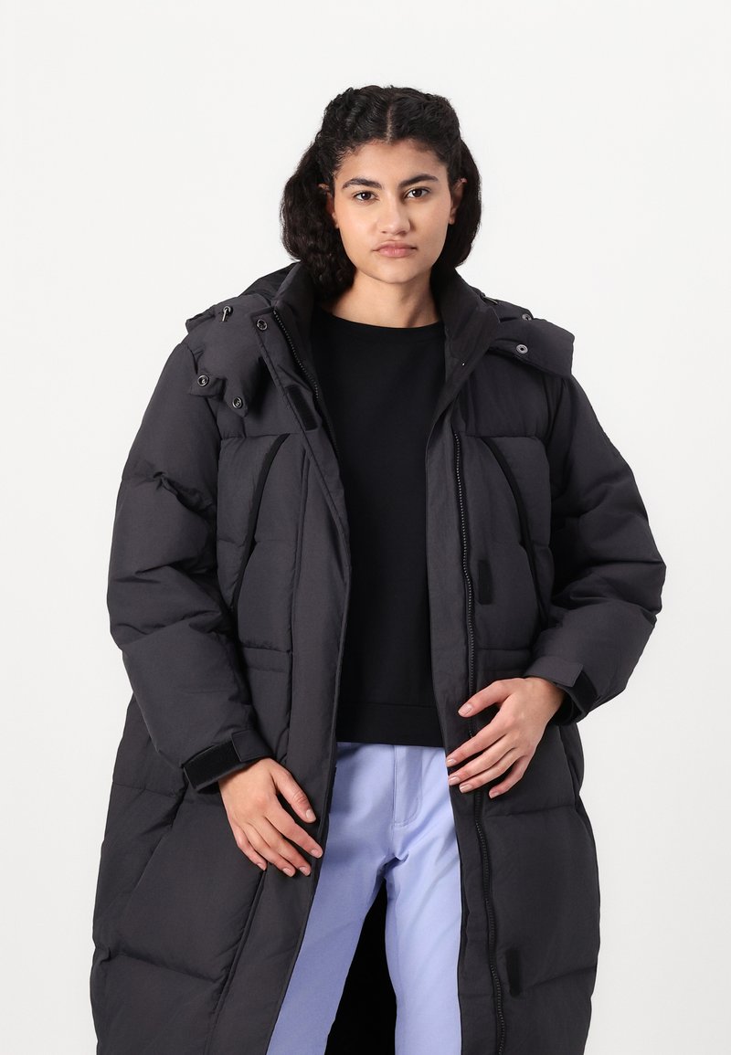 Junge Person, die einen langen schwarzen Steppmantel über einem schwarzen Oberteil und hellblauen Hosen trägt und vor einem schlichten weißen Hintergrund steht.