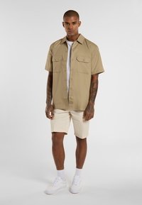 Beige Kurzarmhemd mit zwei Brusttaschen über einem weißen T-Shirt, kombiniert mit cremefarbenen Shorts und weißen Sneakers. Glatter Stofftextur.