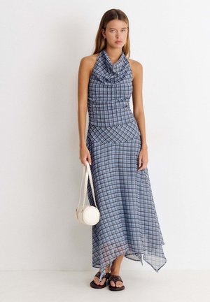 Femme portant une robe à carreaux bleus et noirs à col halter, tenant un petit sac à main rond blanc et portant des sandales noires cloutées.